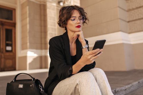 Stylish young woman with short hair wearing cream-colored pants and a black blazer sitting outside on building steps using her phone