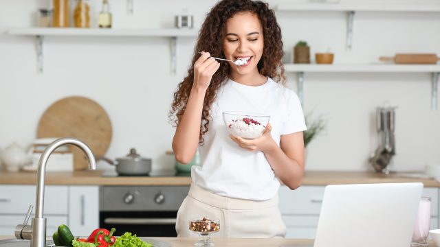 A woman eats cottage cheese, one of the best snacks for weight loss, from a bowl in a sunlit kitchen with white walls.