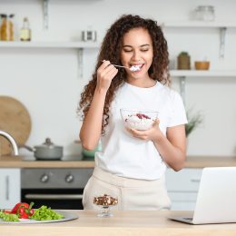 A woman eats cottage cheese, one of the best snacks for weight loss, from a bowl in a sunlit kitchen with white walls.