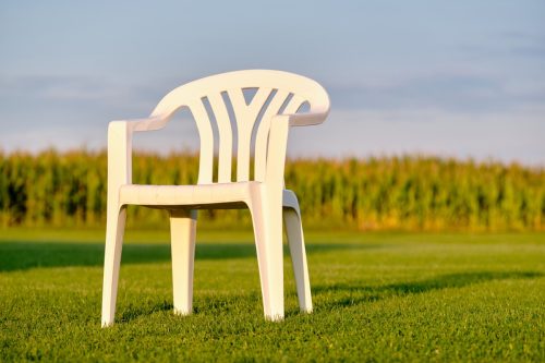 Empy white plastic garden chair standing on a green lawn in front of a cornfield in evening light. Seen in Bavaria, Germany in August.