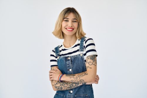 Young woman with tattoos on her arms wearing a black-and-white striped t-shirt and denim overalls against a white background