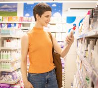 Young woman wearing an orange sleeveless shirt and jeans holds up a bottle of lotion while shopping in a drugstore