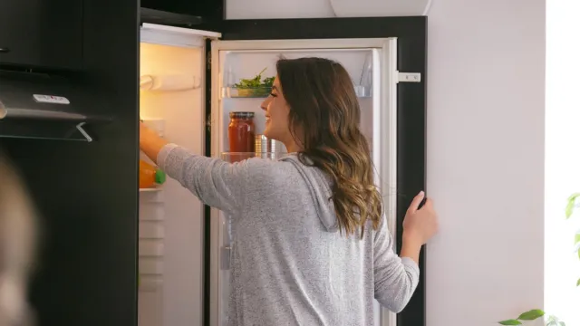Happy woman opening the fridge and taking food from the upper shelf