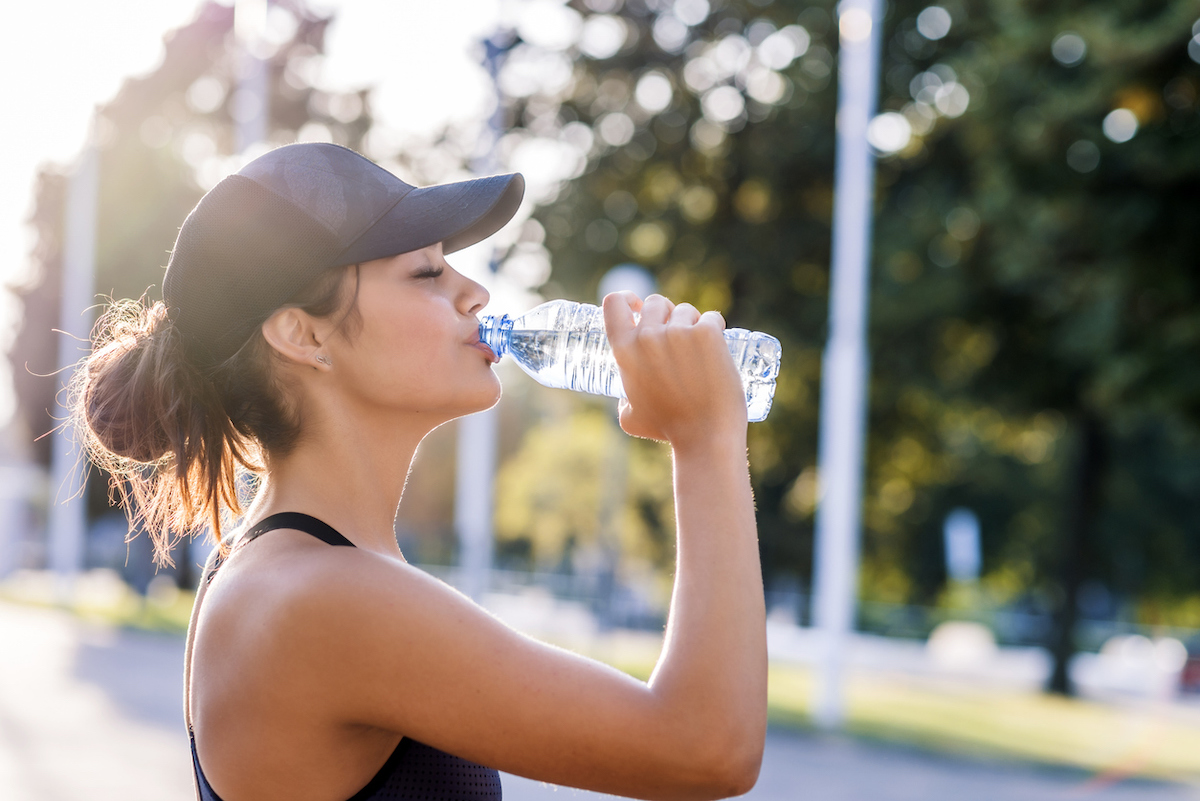 Closeup of a sporty-looking young woman wearing a black baseball cap drinking from a water bottle outside