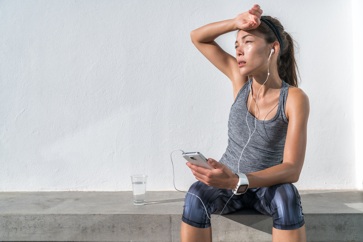 Tired woman taking a break from a run. She's sitting on a concrete ledge listening to music with her hand to her forehead.