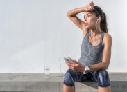 Tired woman taking a break from a run. She's sitting on a concrete ledge listening to music with her hand to her forehead.