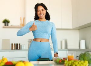 Happy woman in blue workout clothes giving a thumbs up in her kitchen