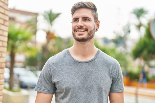 Young man with facial hair wearing a gray t-shirt smiling confident looking to the side at street