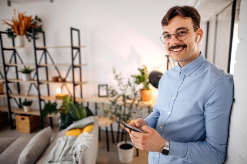 Portrait of a young man with a mustache wearing a light blue shirt on the phone in his living room