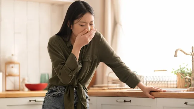 A young woman holding her hand over her mouth, feeling nauseous while standing in her kitchen