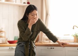 A young woman holding her hand over her mouth, feeling nauseous while standing in her kitchen
