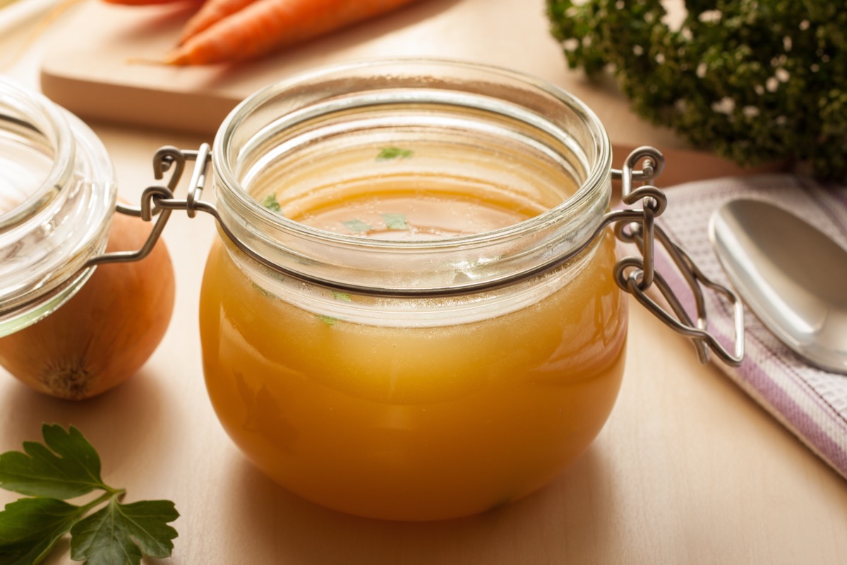 Bone broth in a glass jar, with carrots, onions, and parsley in the background