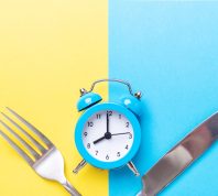 Blue alarm clock, fork, and knife on yellow and blue background. Intermittent fasting concept.