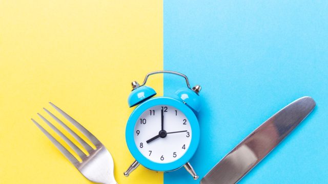 Blue alarm clock, fork, and knife on yellow and blue background. Intermittent fasting concept.