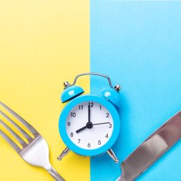 Blue alarm clock, fork, and knife on yellow and blue background. Intermittent fasting concept.