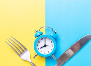 Blue alarm clock, fork, and knife on yellow and blue background. Intermittent fasting concept.
