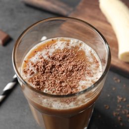 Glass with chocolate protein shake on table, with banana in the background