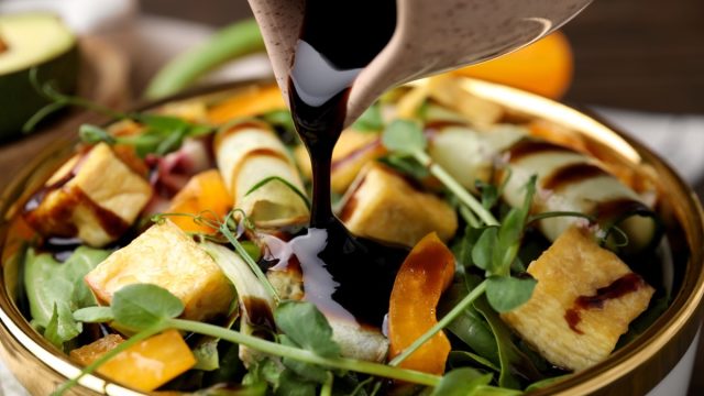 Pouring balsamic vinegar onto delicious salad with tofu and vegetables in bowl, closeup