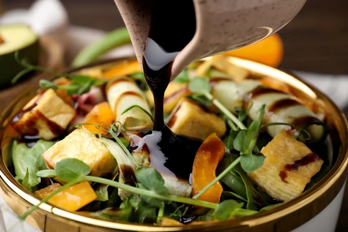 Pouring balsamic vinegar onto delicious salad with tofu and vegetables in bowl, closeup
