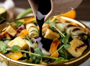Pouring balsamic vinegar onto delicious salad with tofu and vegetables in bowl, closeup