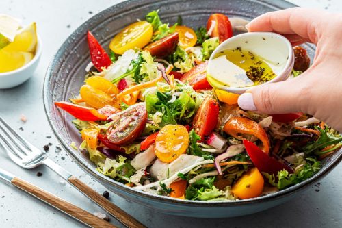 Fresh colorful spring vegetable salad with cherry tomatoes and sweet peppers in the blue bowl. Cook's hand pouring olive oil with herbs (dressing). Healthy organic vegan lunch or snack close up.