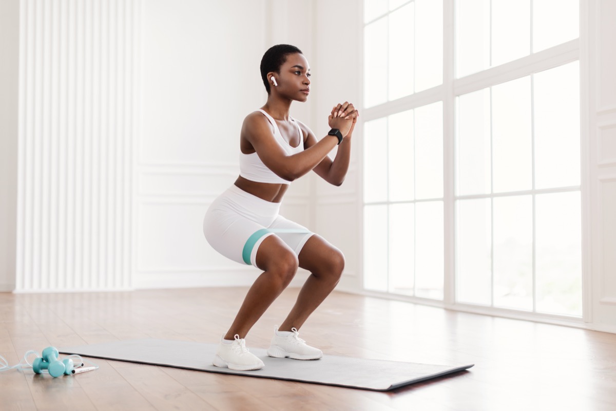 Young woman wearing white shorts and sports bra doing squat workout using resistance bands