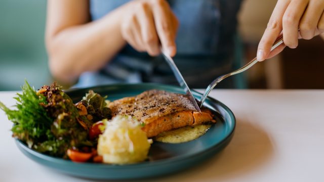 Close up shot of woman eating pan fried salmon with table knife and fork in cafe