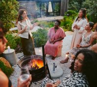 group of smiling women having drinks and talking around a fire pit during an early evening party in the lush garden of a home at dusk