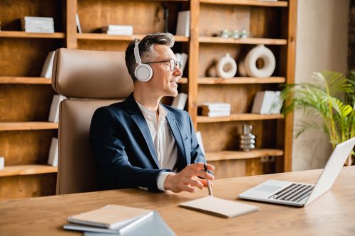 mature middle-aged businessman ceo freelancer boss employee having break after hard-working day in office, relaxing while listening to music radio podcast in headphones at the desk