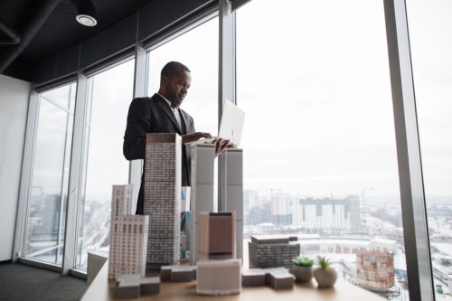 Smiling business man real estate agents in black stylish formal suit standing with laptopnear 3d model skyscrapers of city architecture design at office with panoramic city view.