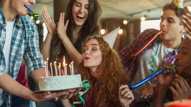 woman in a party hat blowing out birthday candles while surrounded by friends in party hats
