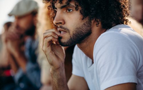 Man looking worried while watching a match at stadium. Man biting nails while watching a live soccer game from stands.