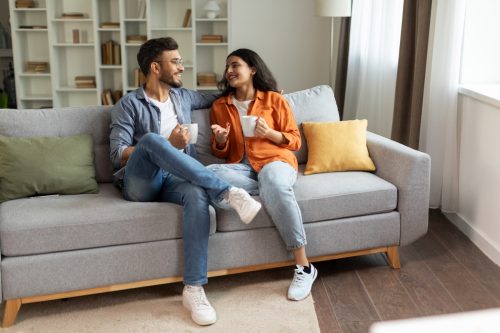 Happy couple relaxing and drinking coffee, sitting on couch at home, talking while resting with hot drinks in cups at modern living room, free space