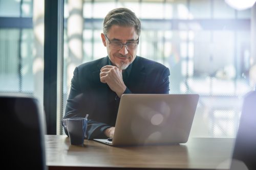 Business portrait - businessman sitting in in office working with laptop computer. Mature age, middle age, mid adult man in 50s with happy confident smile. Copy space.