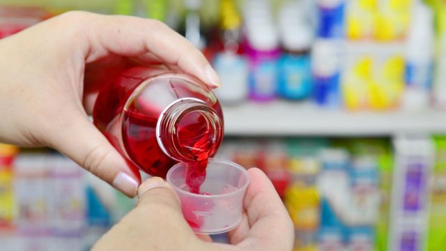 Pouring red cough and cold liquid medicine into measuring cup on pharmacy drugstore background.