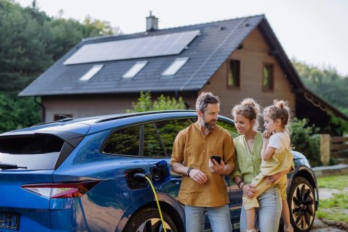 Parents with little girl standing in front of their blue electric vehicle with a house with solar panels on the roof in the background