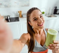selective focus of positive girl holding green smoothie and looking at camera