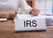 Close-up Of Nameplate With IRS Title Kept On Desk In Front Of Businesswoman Working