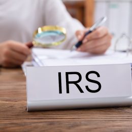 Close-up Of Nameplate With IRS Title Kept On Desk In Front Of Businesswoman Working