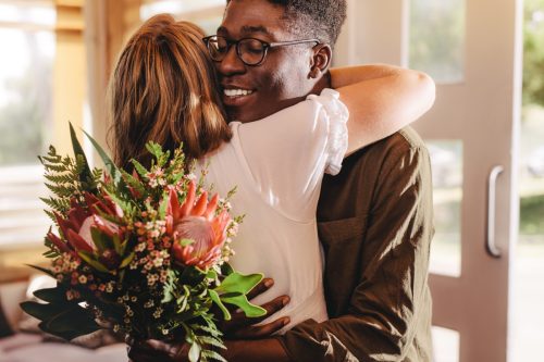 Young man hugging his girlfriend standing in cafe. Young guy expressing his love for his lady giving flowers and a warm hug during a coffee shop date.
