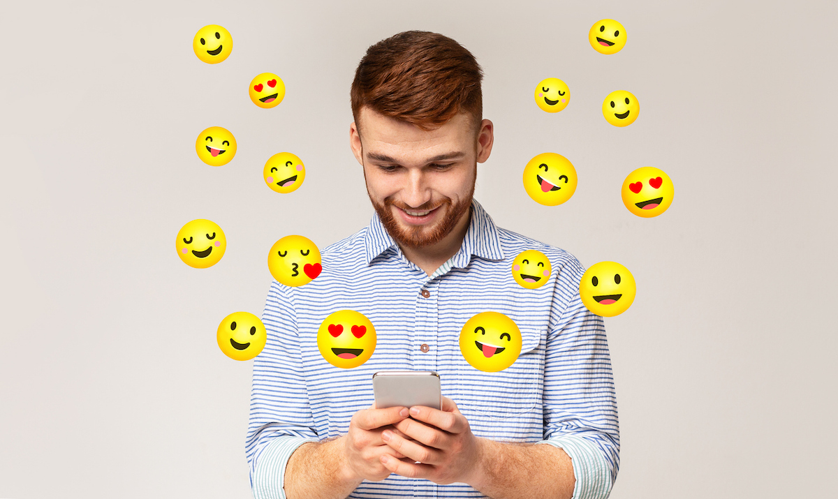 Happy young man texting with flirty emojis surrounding him on gray background
