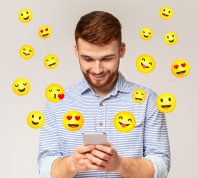 Happy young man texting with flirty emojis surrounding him on gray background