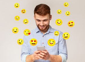 Happy young man texting with flirty emojis surrounding him on gray background