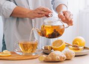 Cropped shot of a person pouring lemon-ginger tea from a glass teapot into a glass mug on a table surrounded by lemons and ginger