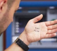 closeup of a man looking at the palm of his hand with "pin 1234" written on it, standing in front of an ATM.