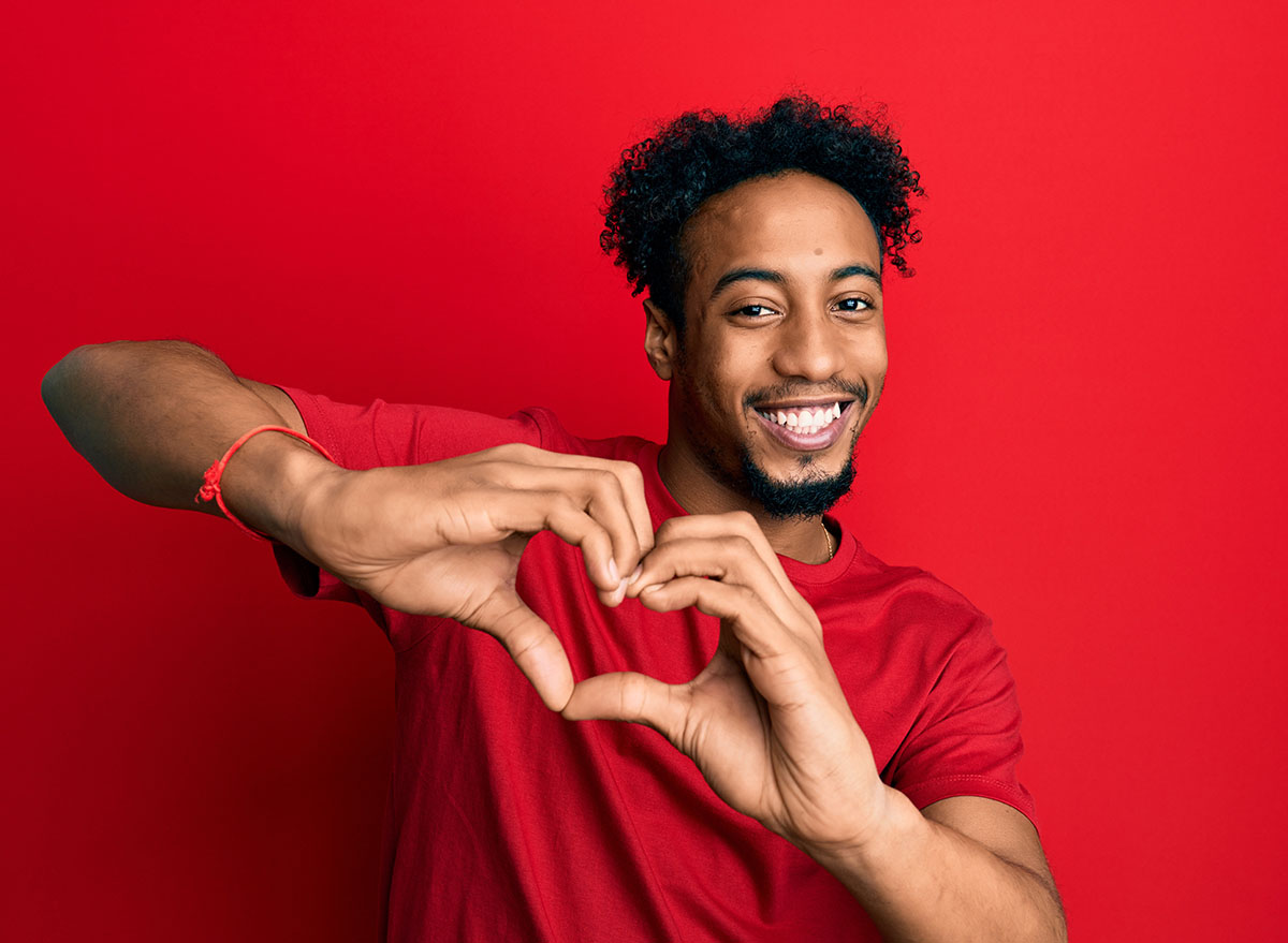 Young african american man with beard wearing casual red t shirt smiling in love doing heart symbol shape with hands. romantic concept.