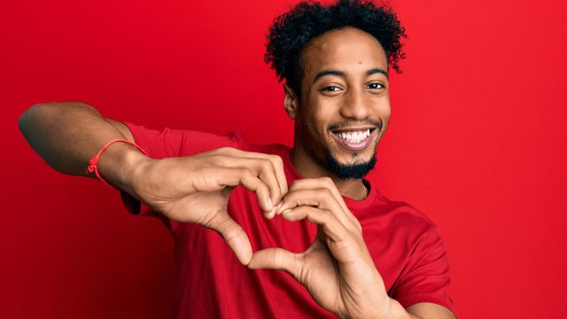 Young african american man with beard wearing casual red t shirt smiling in love doing heart symbol shape with hands. romantic concept.