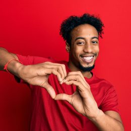 Young african american man with beard wearing casual red t shirt smiling in love doing heart symbol shape with hands. romantic concept.