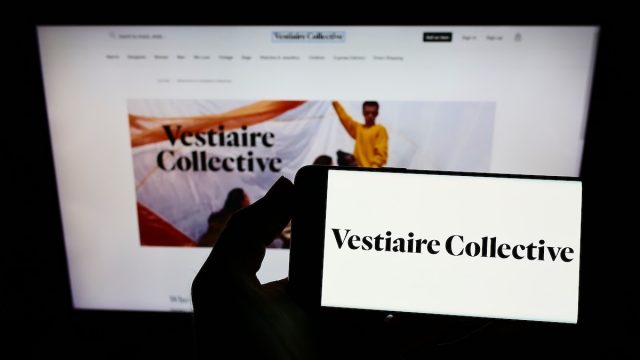 Person holding smartphone with logo of French e-commerce company Vestiaire Collective SA on screen in front of website.