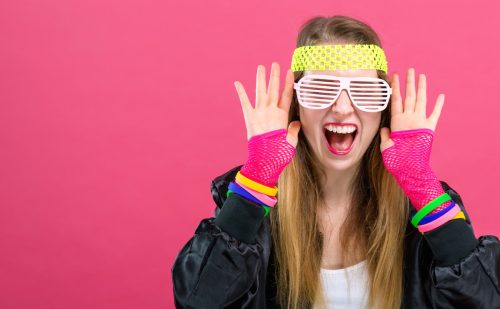 Woman in 1980's fashion theme on a pink background. She's wearing a black jacket, pink fingerless gloves, a yellow headband, and white glasses
